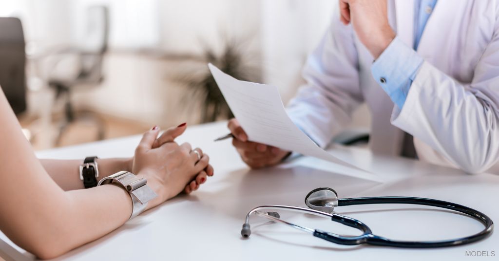 Woman's hands on table with doctor in a white coat holding a piece of paper. (MODELS)